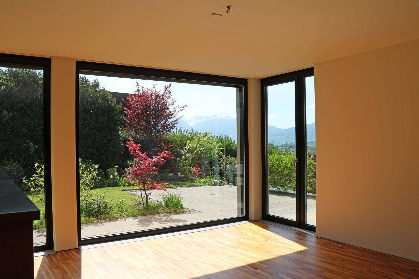 Heller Wohnbereich mit Holzparkettboden und raumhohen Fenstern, die einen Garten- und Bergblick bieten.