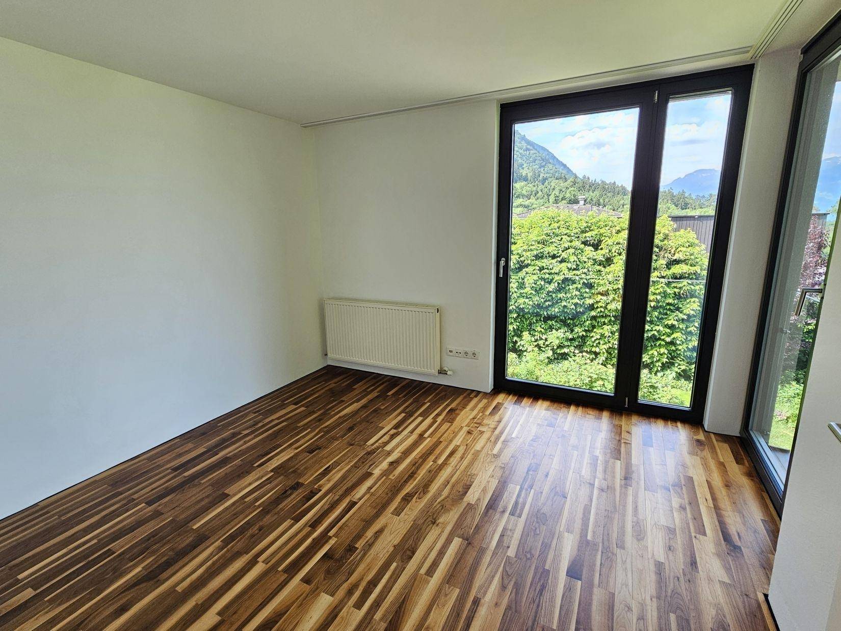 Helles Zimmer mit Holzparkettboden, Heizkörper und großen Fenstern mit Bergblick.