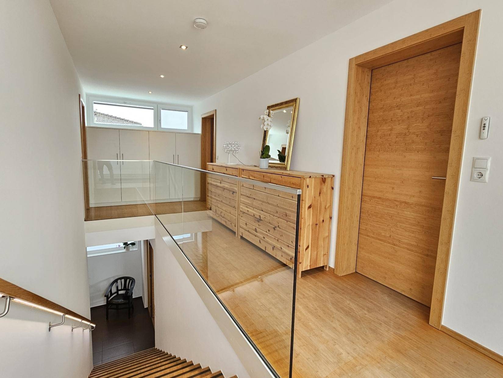 Heller Flur mit Holztreppe, Glasgeländer und einem Holzschrank mit Spiegel.