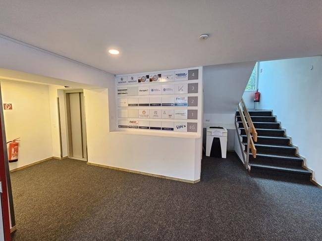 Heller Empfangsbereich mit Teppichboden, Informationswand, Treppe und einem Aufzug.