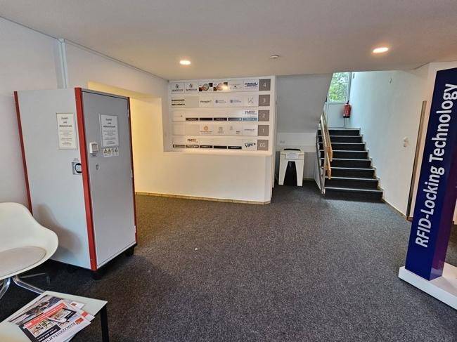 Empfangsbereich mit Teppichboden, Informationswand, Schließfächern und Treppenaufgang.