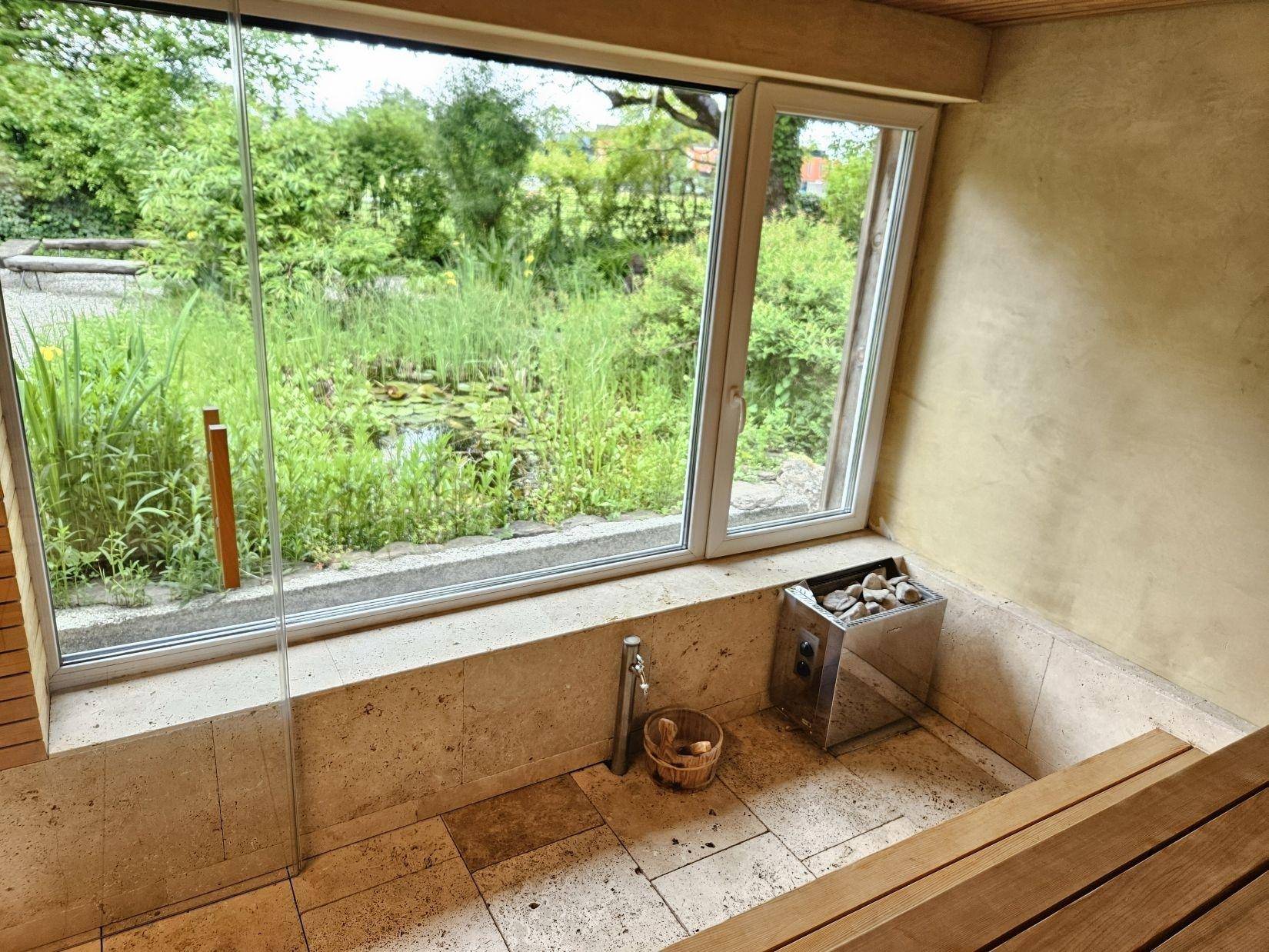 Sauna mit Blick ins Grüne durch große Fenster, ausgestattet mit einem Saunaofen und Holzbänken.