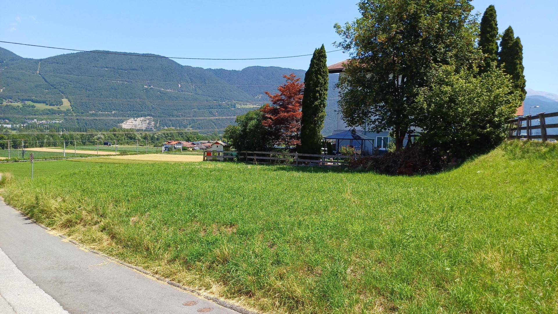 Weitläufige grüne Wiese mit Blick auf die Ferne, umgeben von Bäumen und Bergen am Horizont.