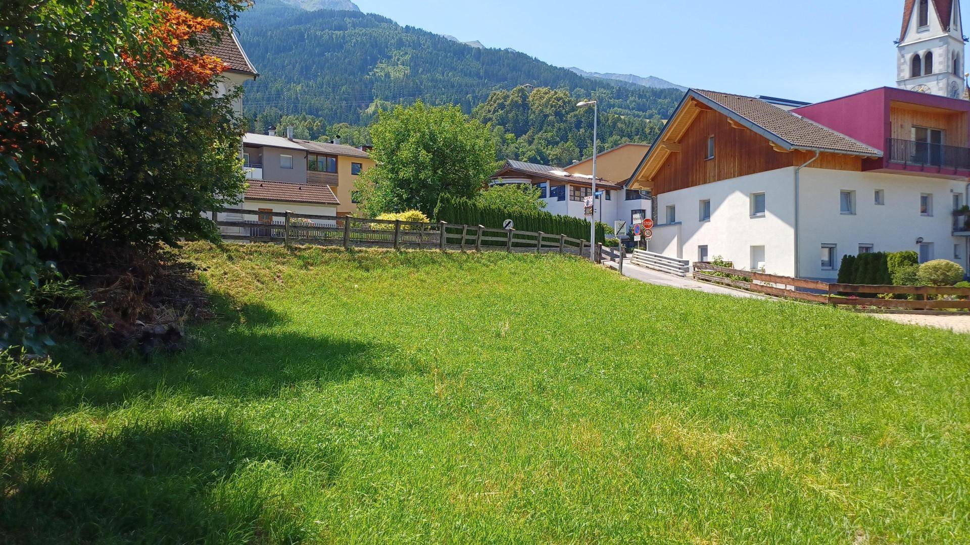 Grüne Wiese mit Blick auf die umliegenden Häuser und bewaldeten Hügel unter blauem Himmel.