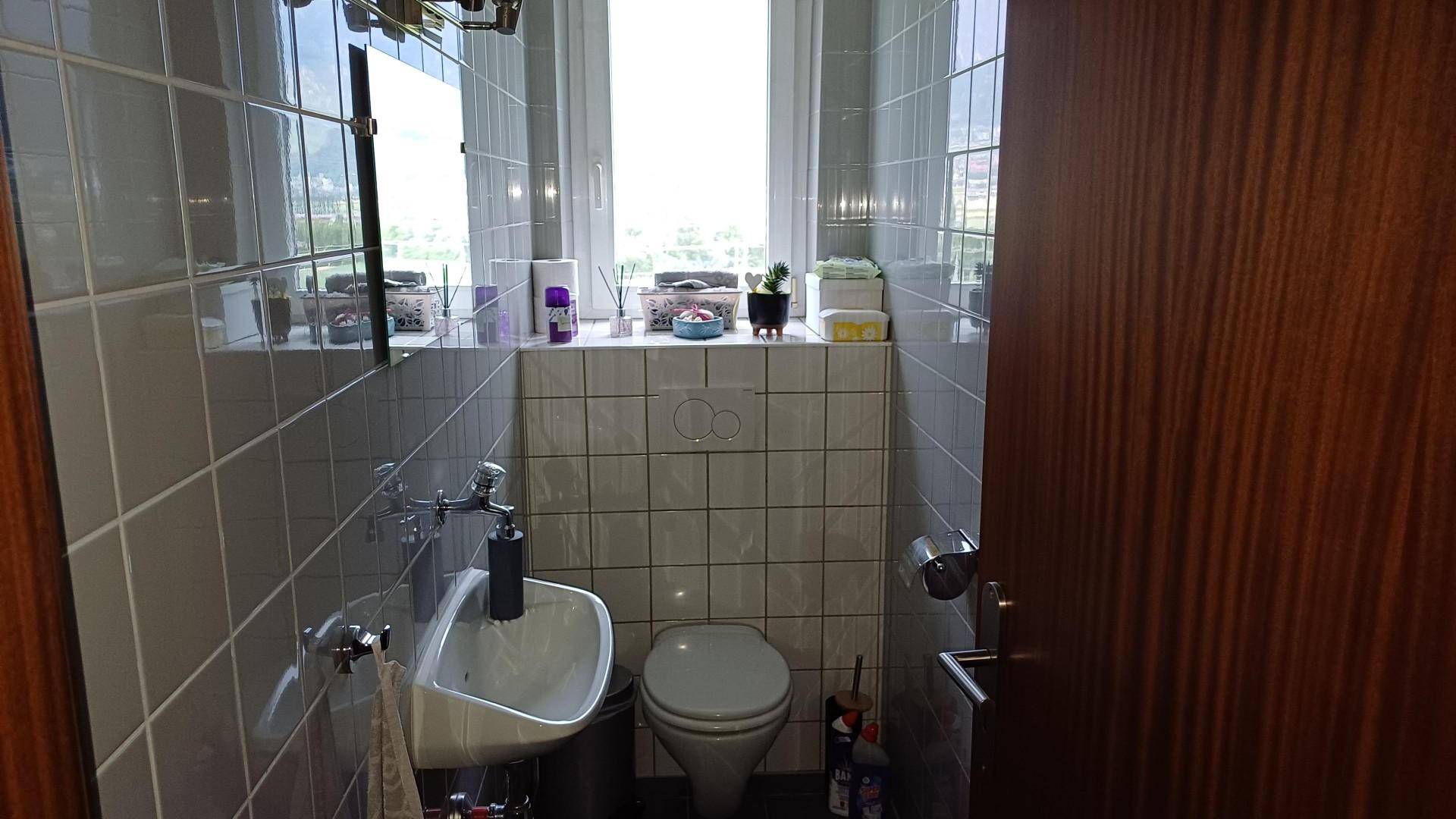 Kleines Gäste-WC mit Fenster, Wand-WC und Waschbecken, ausgestattet mit hellen Fliesen.