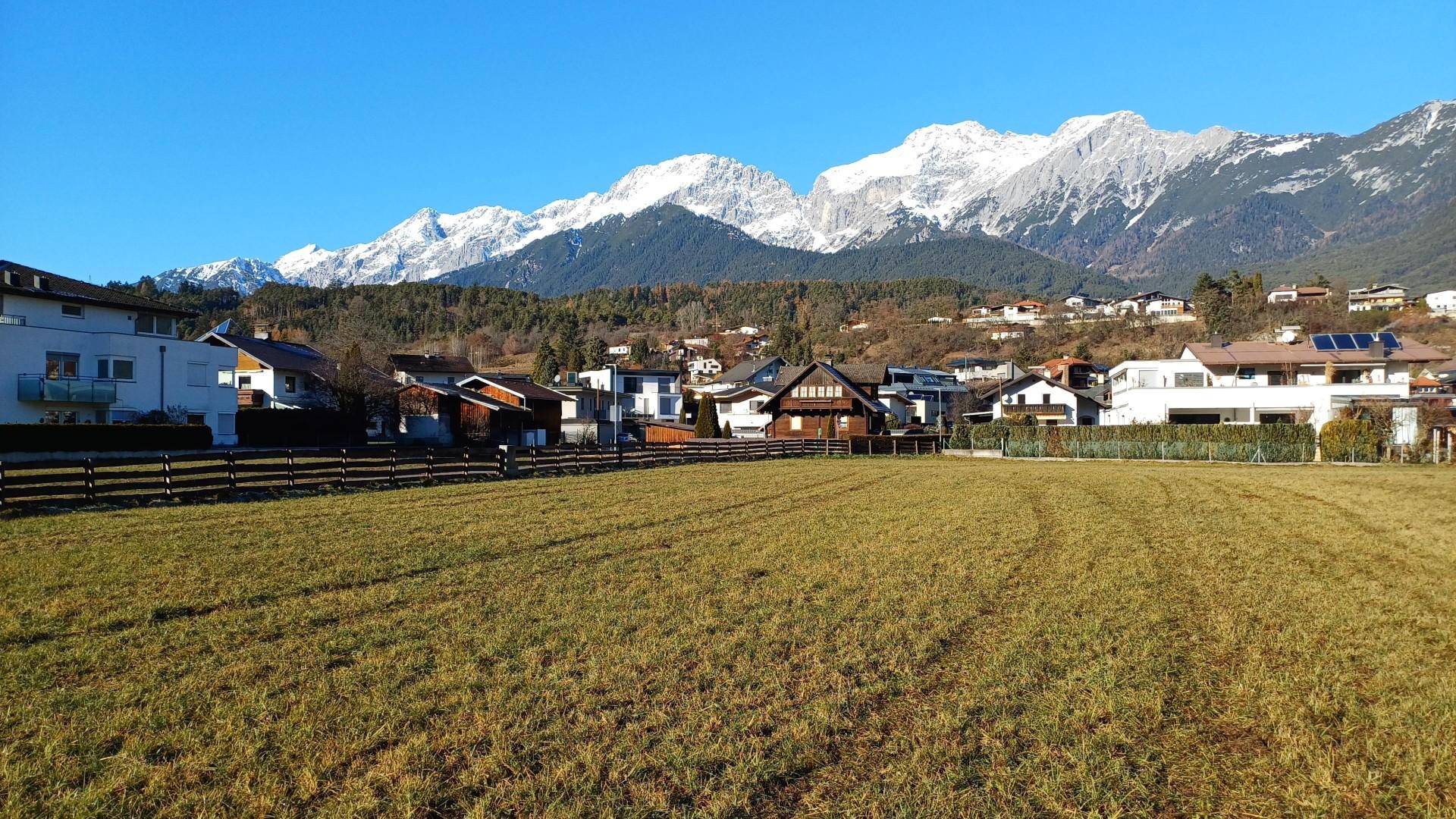 Panoramablick über ein weites Feld auf eine Wohnsiedlung vor einer beeindruckenden Bergkulisse.