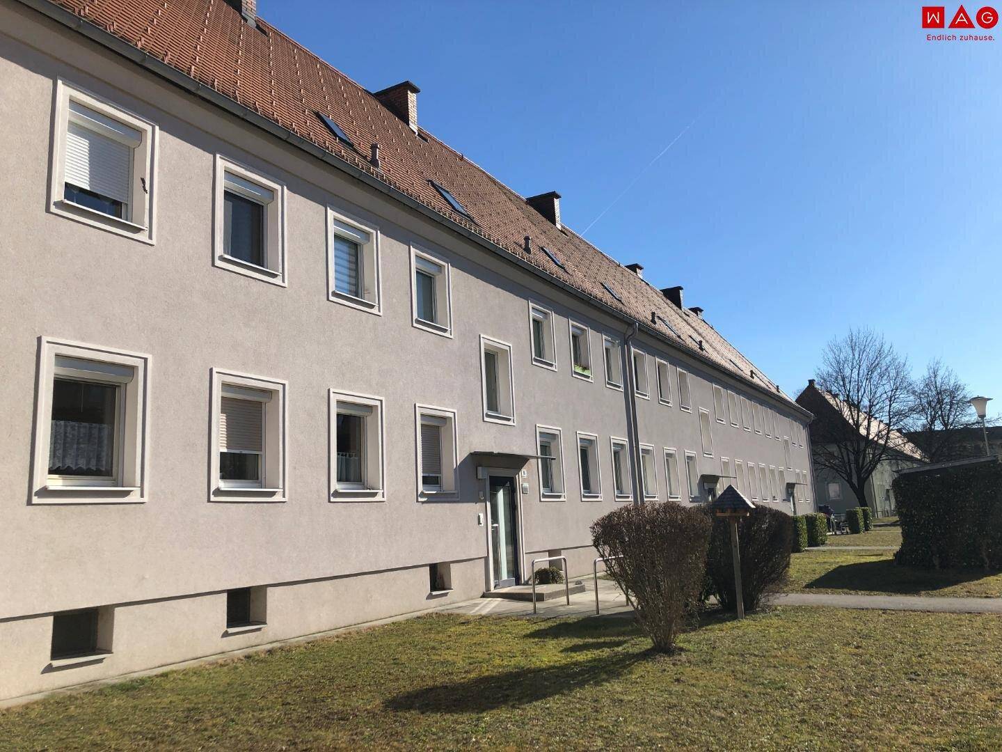 Lange Fassade eines Wohngebäudes mit vielen Fenstern und rotem Ziegeldach an einem sonnigen Tag.