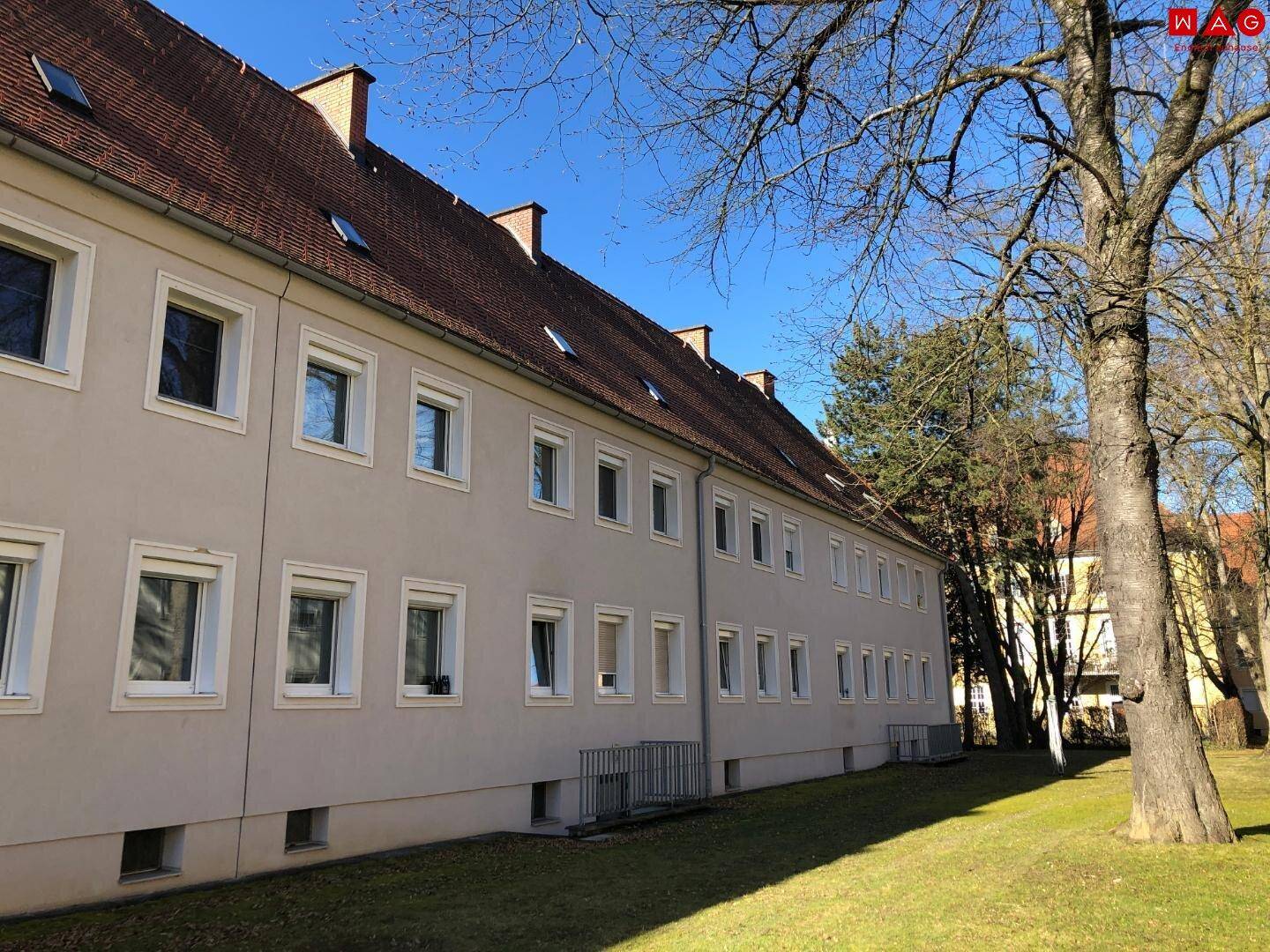 Seitenansicht eines Wohngebäudes mit mehreren Fenstern, rotem Dach und Bäumen im Vordergrund.