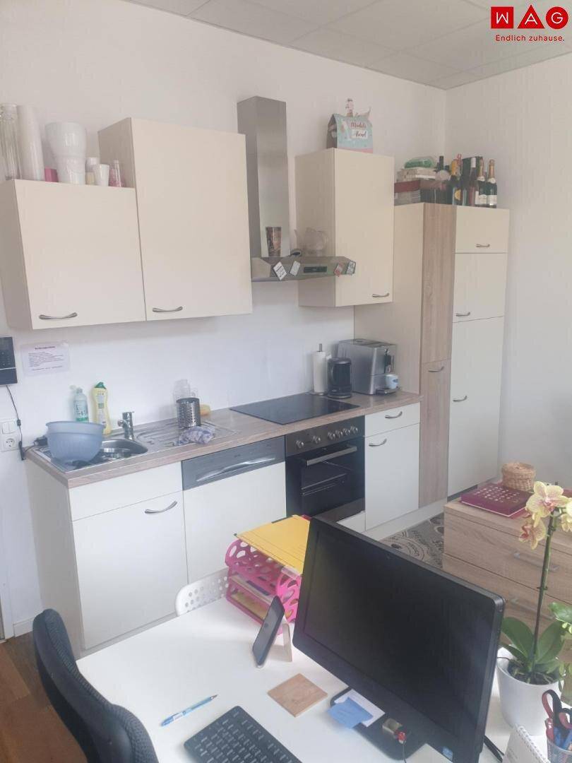 Kleine Einbauküche mit weißen Schränken, Kochfeld und Backofen in einem Büroraum.
