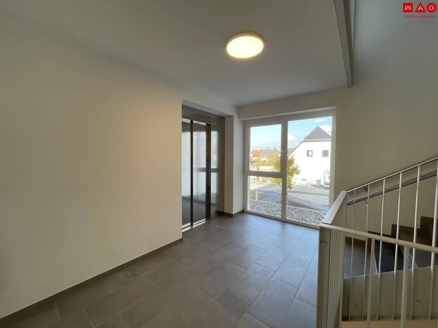 Heller Eingangsbereich des Gebäudes mit Aufzug, Treppenhaus und großen Fenstern, die Tageslicht spenden.