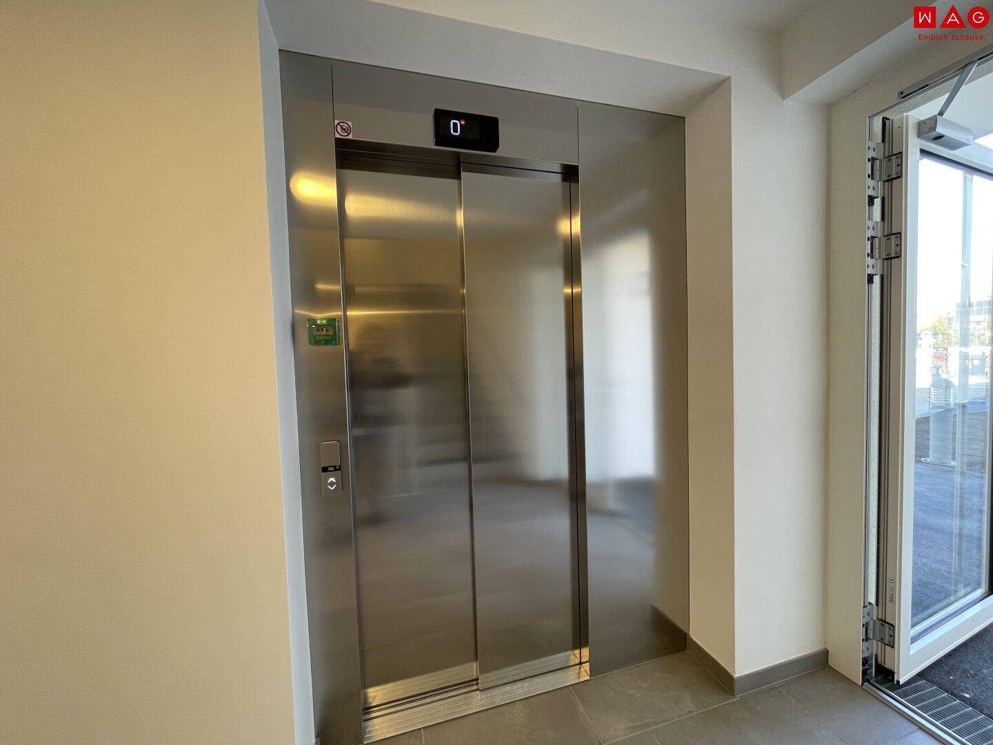 Moderner Aufzug mit Edelstahltüren im hellen Treppenhaus eines Mehrfamilienhauses.