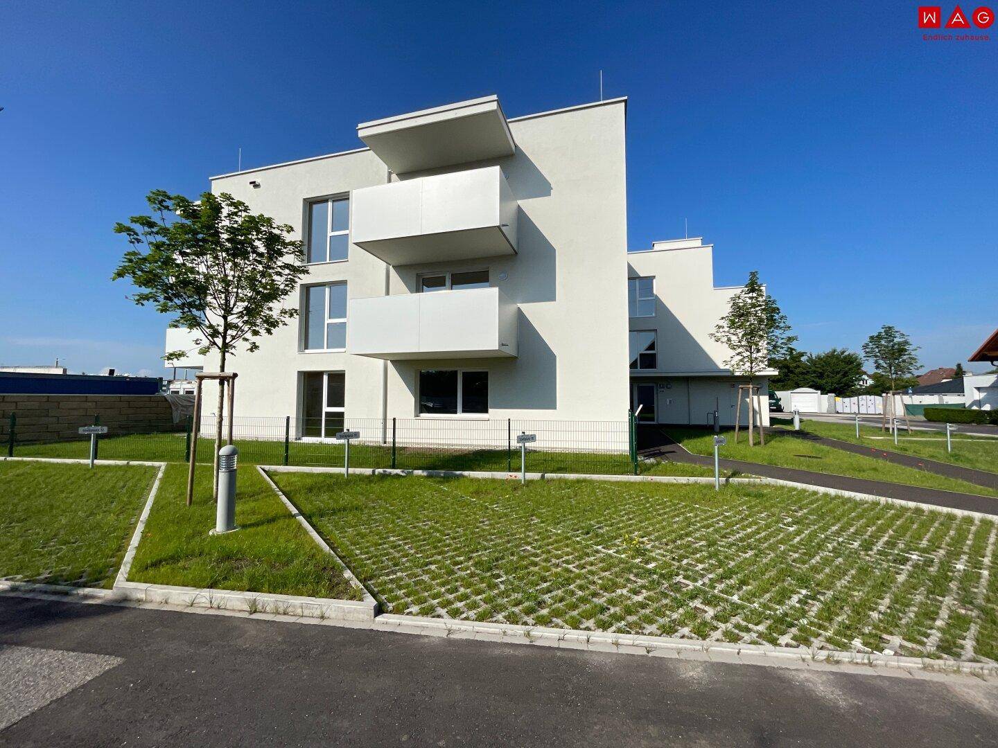 Modernes Mehrfamilienhaus mit klaren Linien, Balkonen und begrünten Außenanlagen unter blauem Himmel.