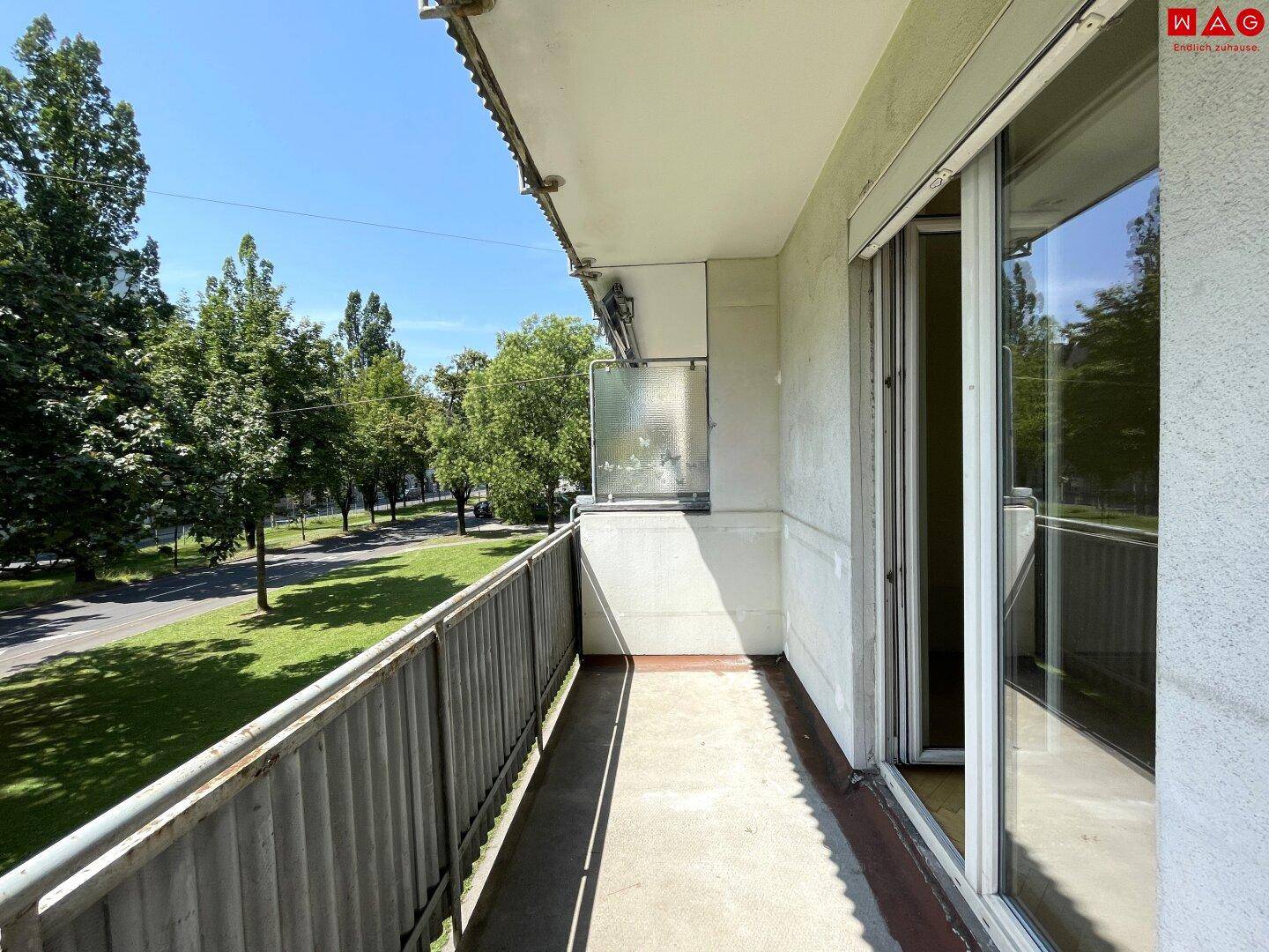 Sonniger Balkon mit Metallgeländer und Blick auf eine von Bäumen gesäumte Straße.