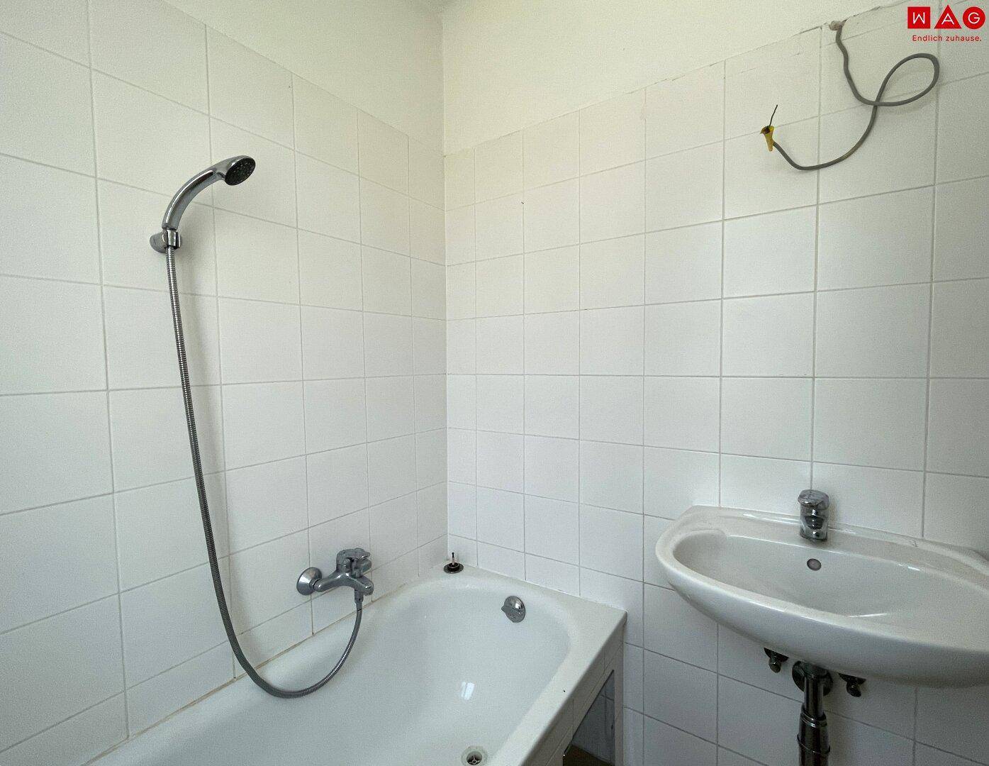Einfaches Badezimmer mit Badewanne, Duschkopf und Waschbecken, komplett gefliest.