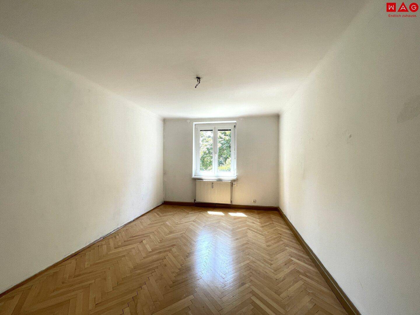 Leeres Zimmer mit Fischgrätparkett und einem großen Fenster, das eine grüne Aussicht bietet.