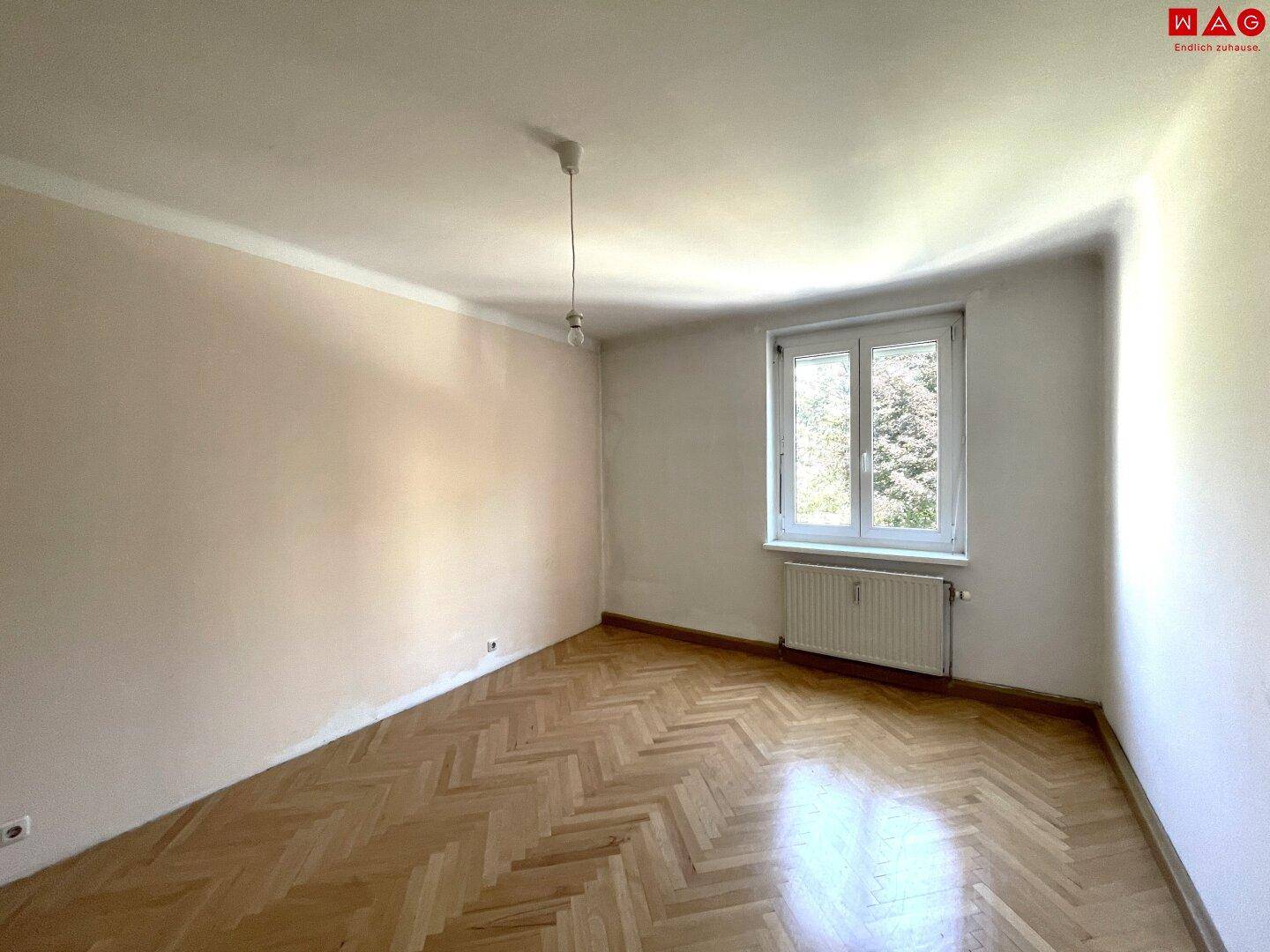 Geräumiges Zimmer mit klassischem Fischgrätparkett und einem Fenster, das viel Tageslicht hereinlässt.