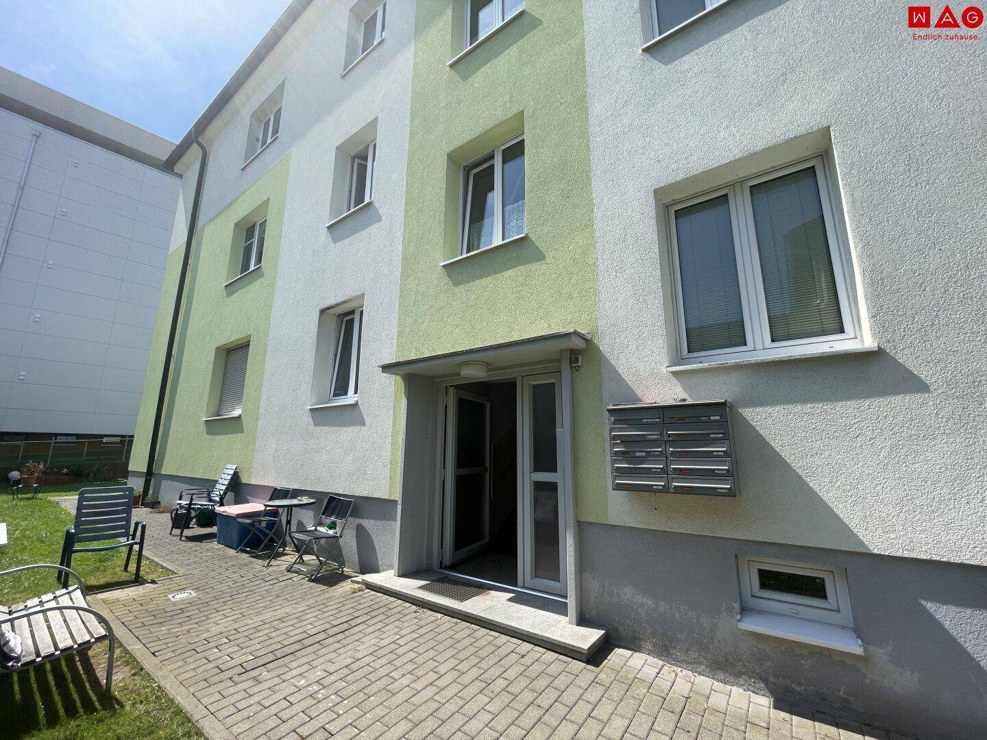 Eingangsbereich eines Wohnhauses mit Briefkästen, gepflastertem Zugang und angrenzender Grünfläche.