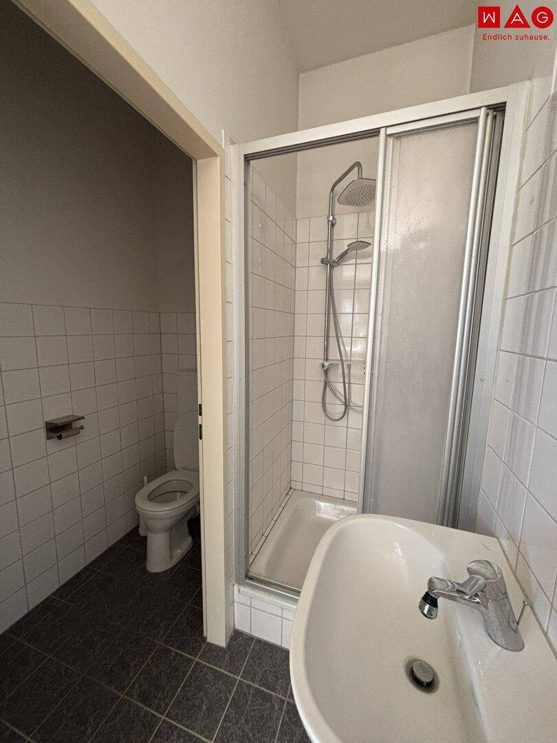 Funktionales Badezimmer mit Duschkabine, Waschbecken und Toilette, gefliest in Weiß und Grau.