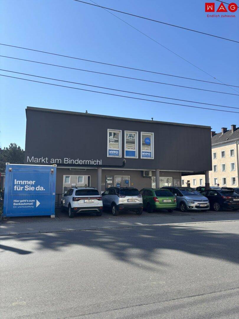 Gebäudeansicht mit geparkten Autos und einem blauen Container, der auf einen Markt hinweist.