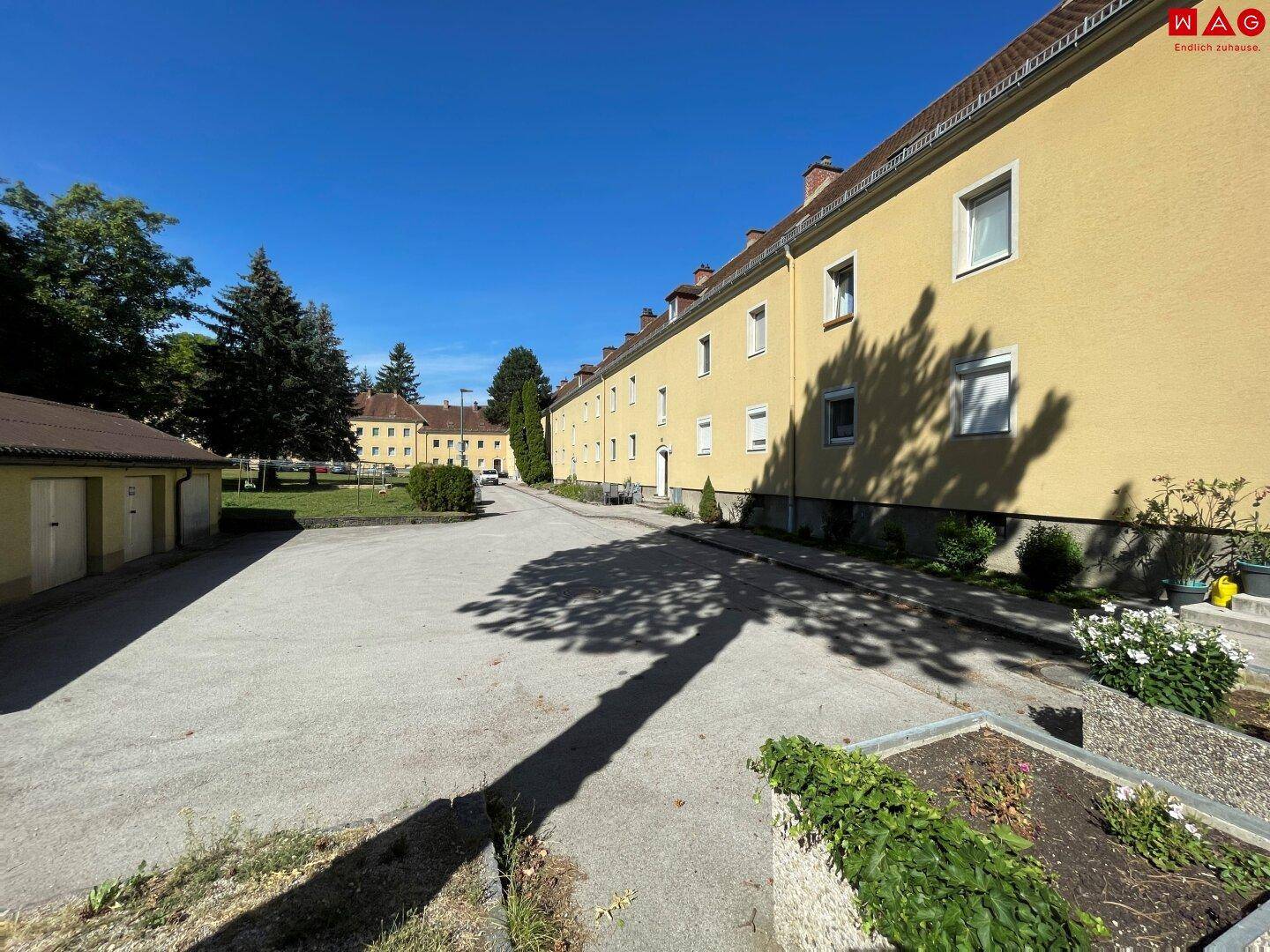 Langes Wohngebäude mit gelber Fassade und gepflastertem Bereich an einem sonnigen Tag.
