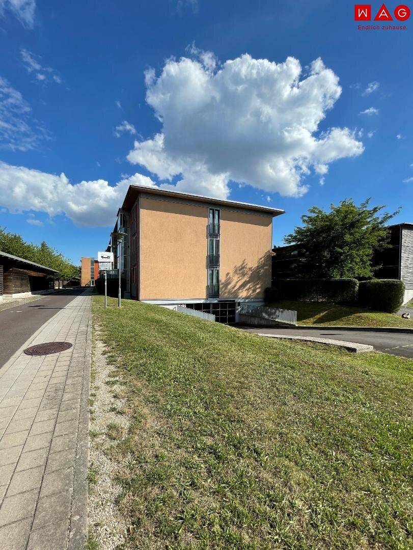Modernes Wohngebäude mit orangefarbener Fassade, umgeben von Grünflächen und einem Gehweg unter blauem Himmel.