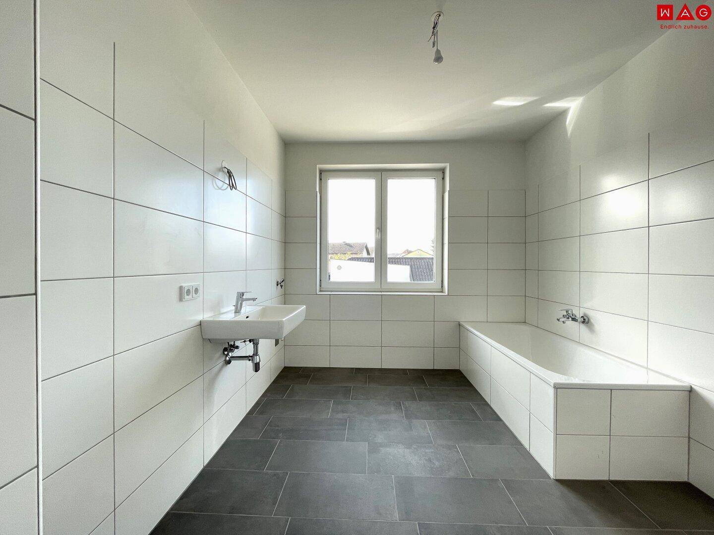 Geräumiges Badezimmer mit Badewanne, Waschbecken und großen weißen Fliesen an Wänden und Boden.