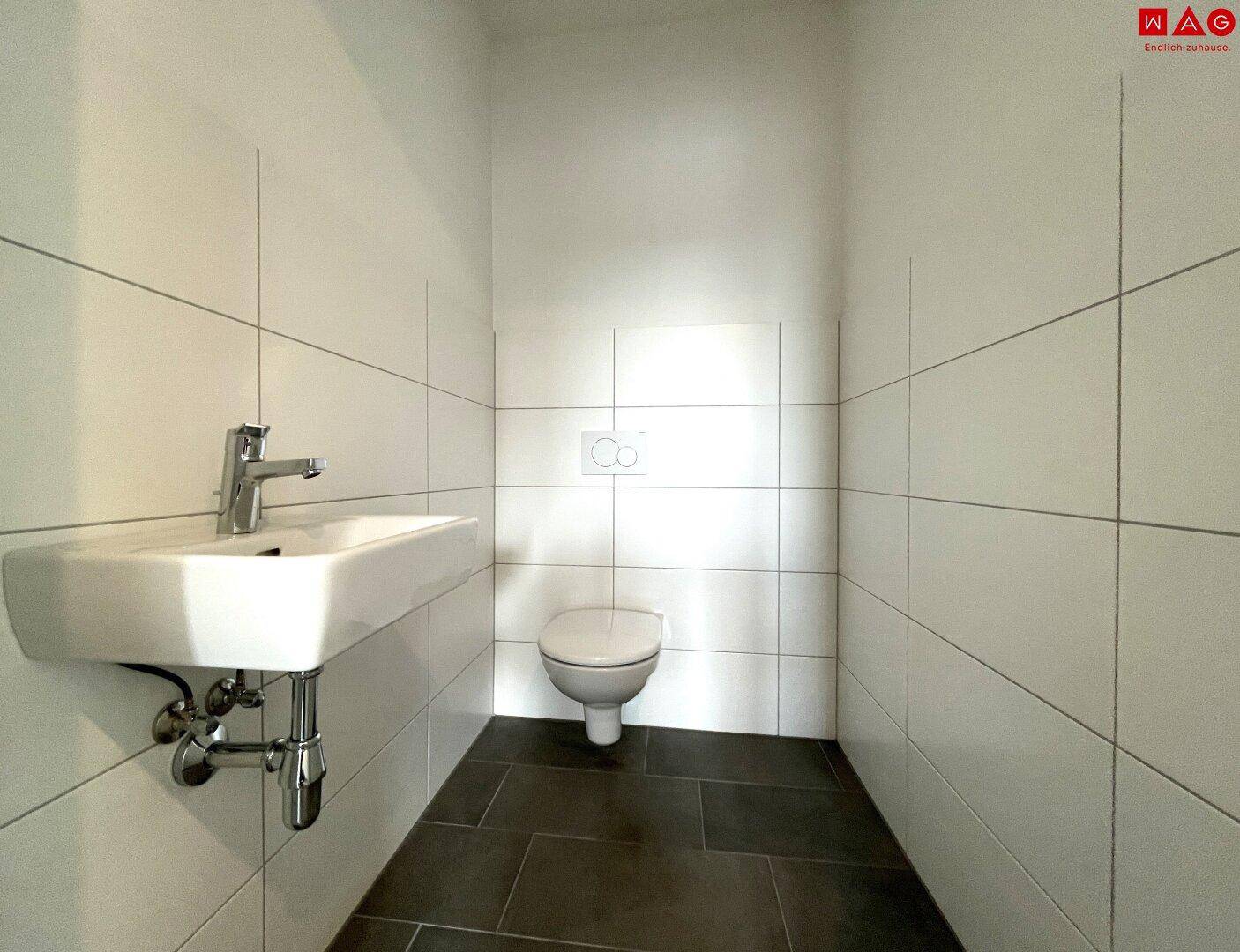 Helles Gäste-WC mit wandhängender Toilette, Waschbecken und weißen Fliesen an Wänden und Boden.