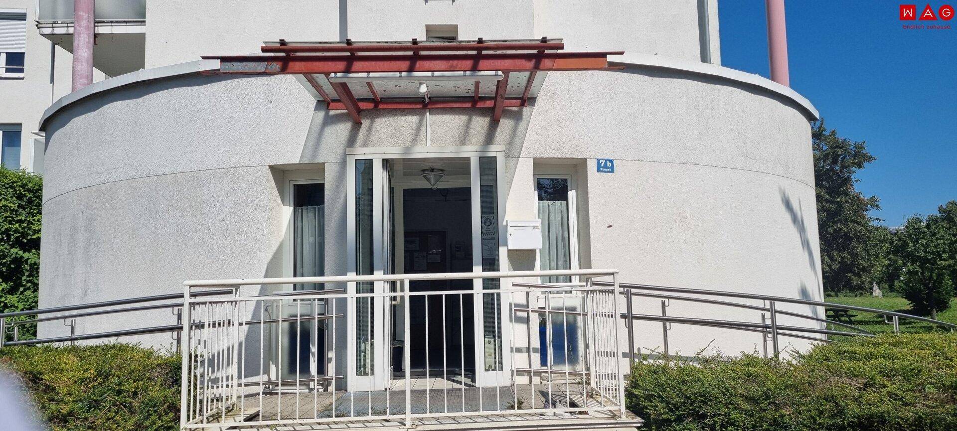 Eingangsbereich eines modernen Gebäudes mit barrierefreier Rampe und gepflegter Grünanlage.