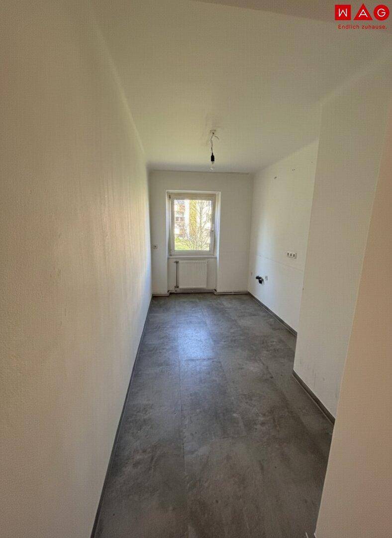 Leerer, schmaler Raum mit Betonboden und einem Fenster, vorbereitet für eine Kücheninstallation.
