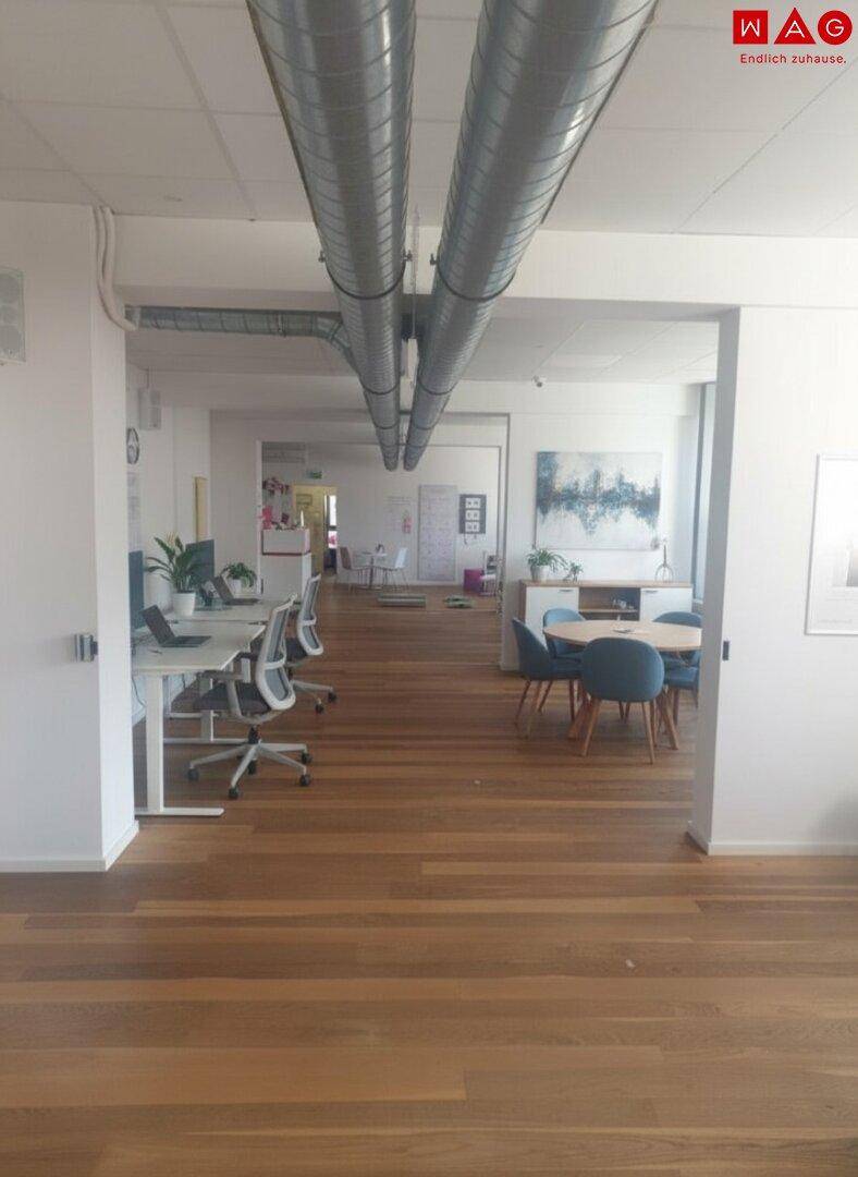 Offener Bürobereich mit Holzboden, Schreibtischen und einem kleinen Besprechungsbereich unter sichtbaren Rohren.