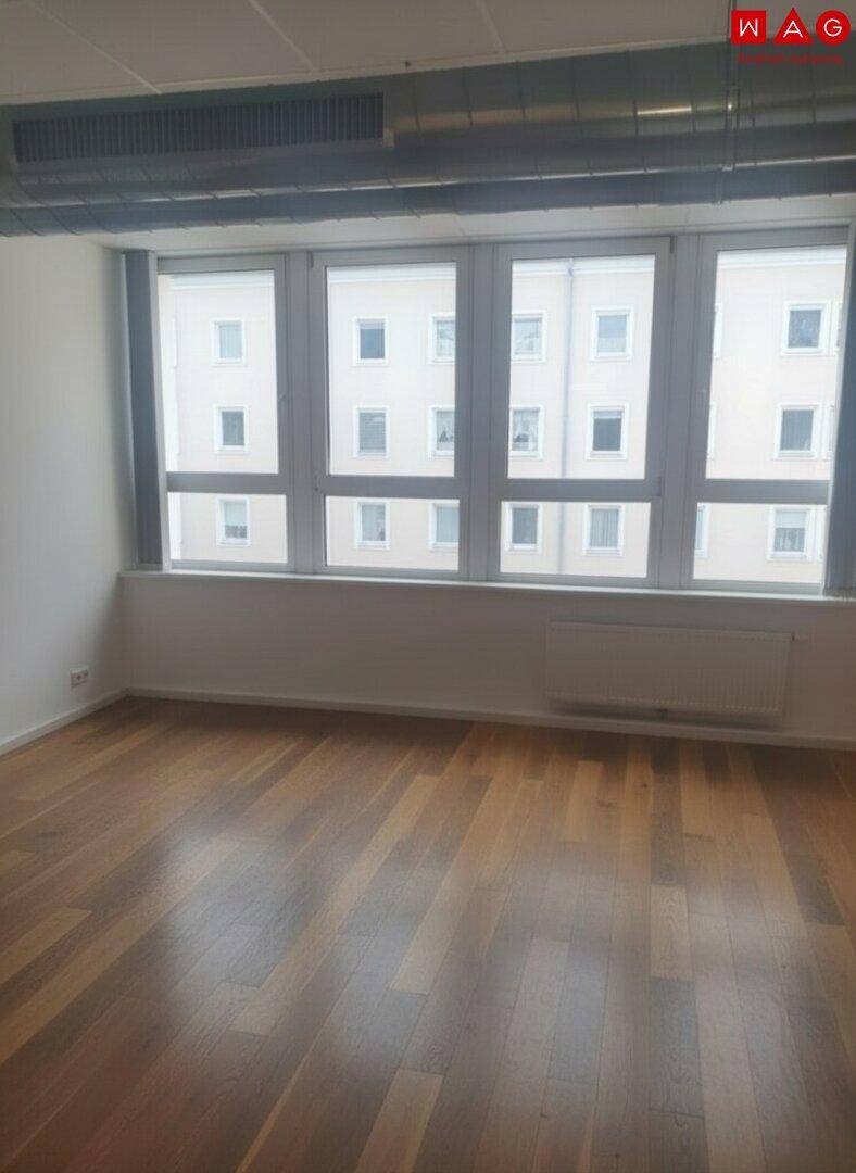 Leerer Büroraum mit Holzboden und großen Fenstern, die viel Tageslicht hereinlassen.