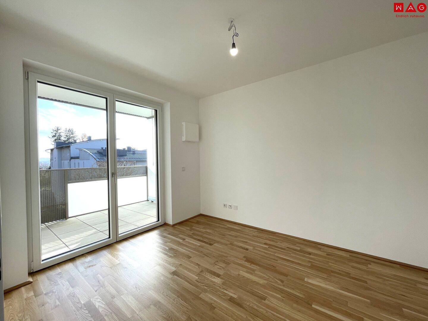 Lichtdurchflutetes Zimmer mit Parkettboden und großem Balkonfenster mit Blick auf die Umgebung.