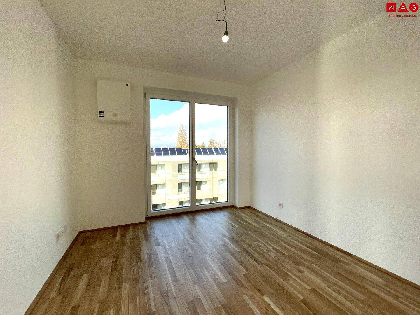 Unmöbliertes Zimmer mit Holzboden und großem Fenster, das viel Tageslicht hereinlässt.