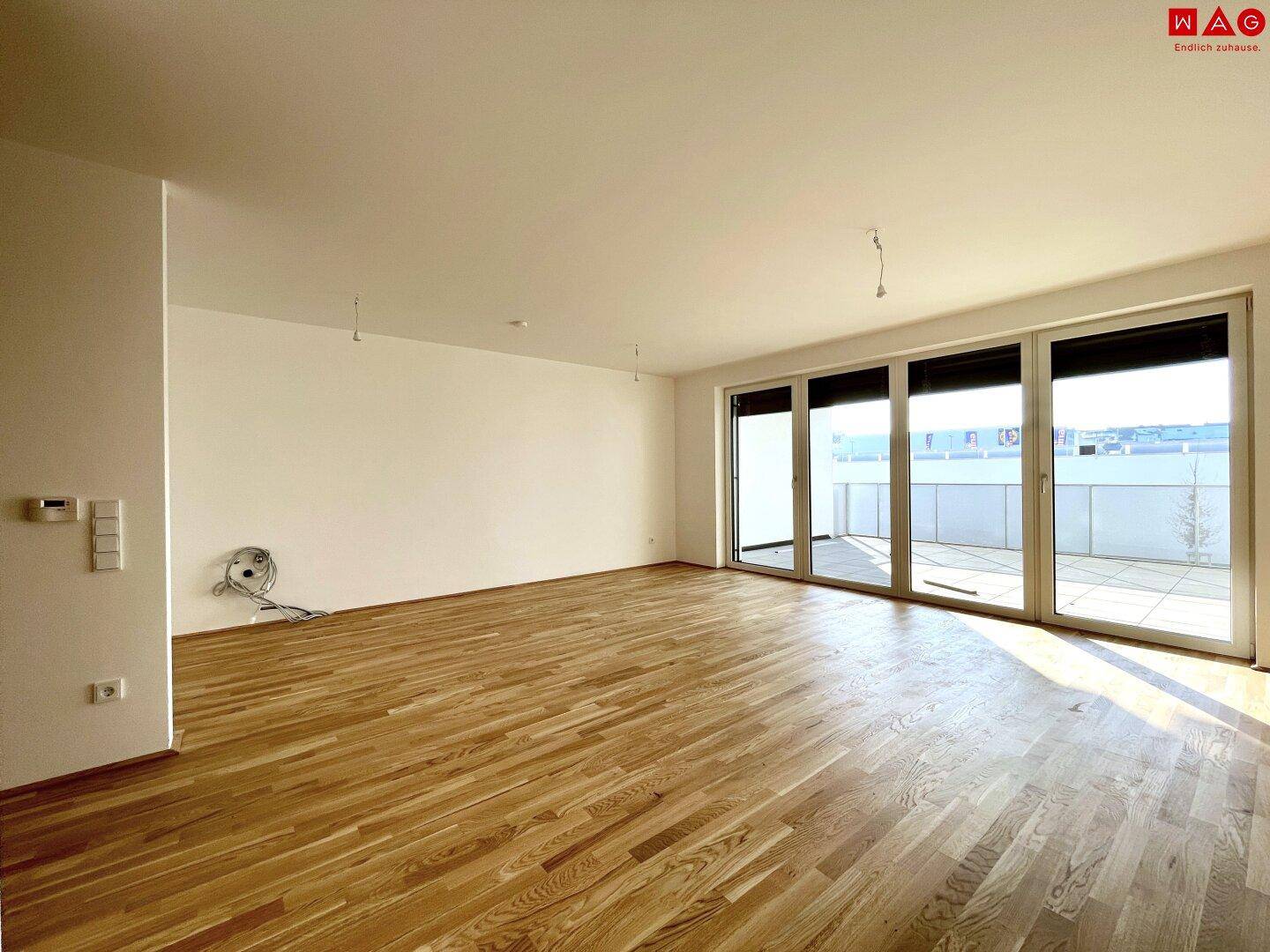 Geräumiges, leeres Wohnzimmer mit hellem Holzboden und großen Fenstern, die auf einen Balkon führen.