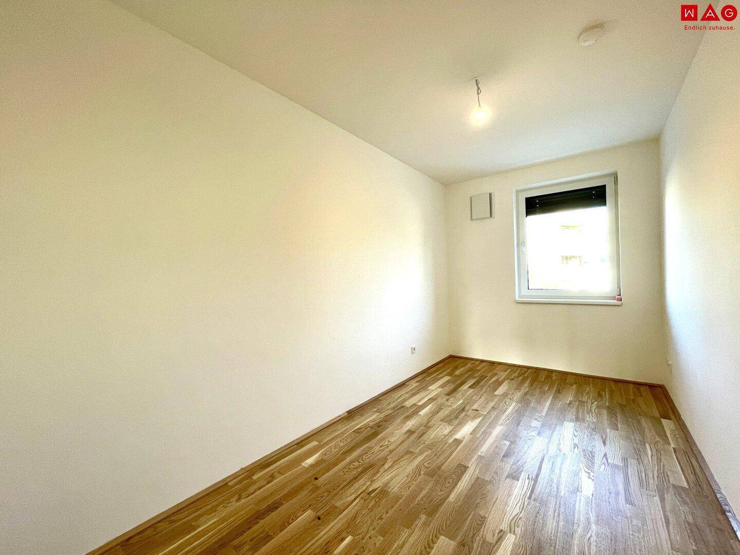Leeres Schlafzimmer mit Holzboden und einem Fenster, das viel Tageslicht hereinlässt.