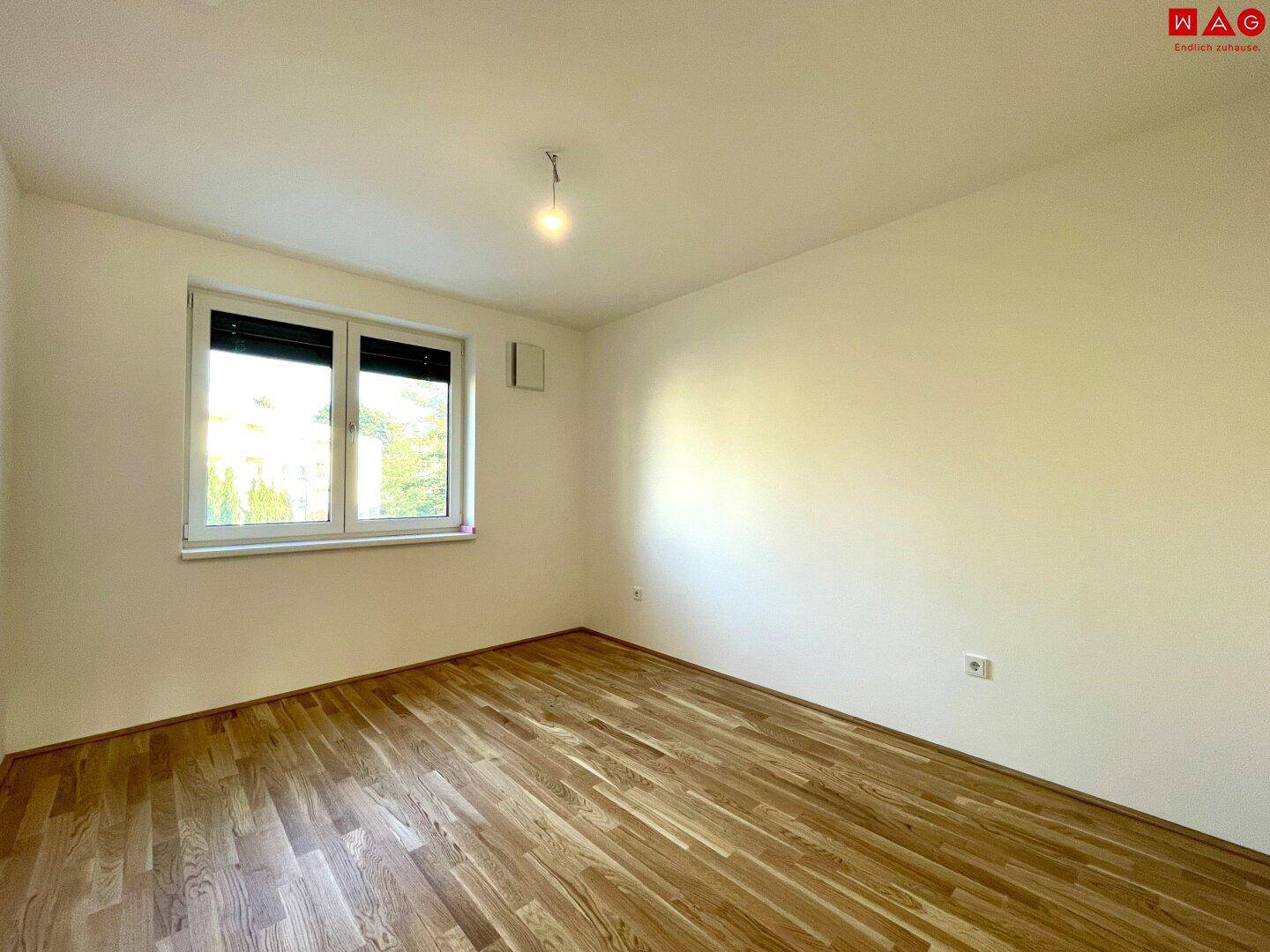 Leeres Schlafzimmer mit Holzboden und einem großen Fenster, das den Raum erhellt.