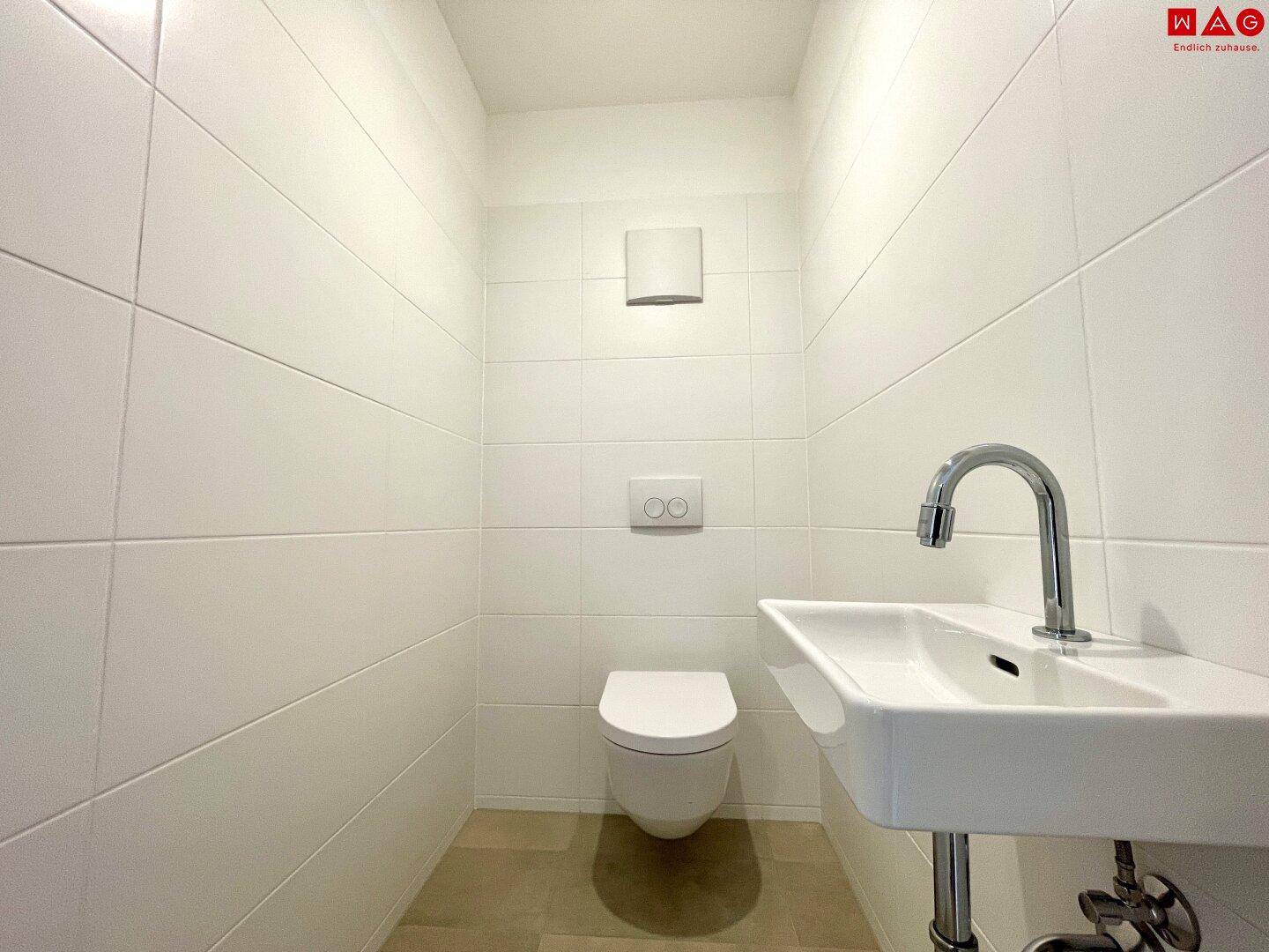 Helles Gäste-WC mit wandhängender Toilette und kleinem Waschbecken, vollständig gefliest.