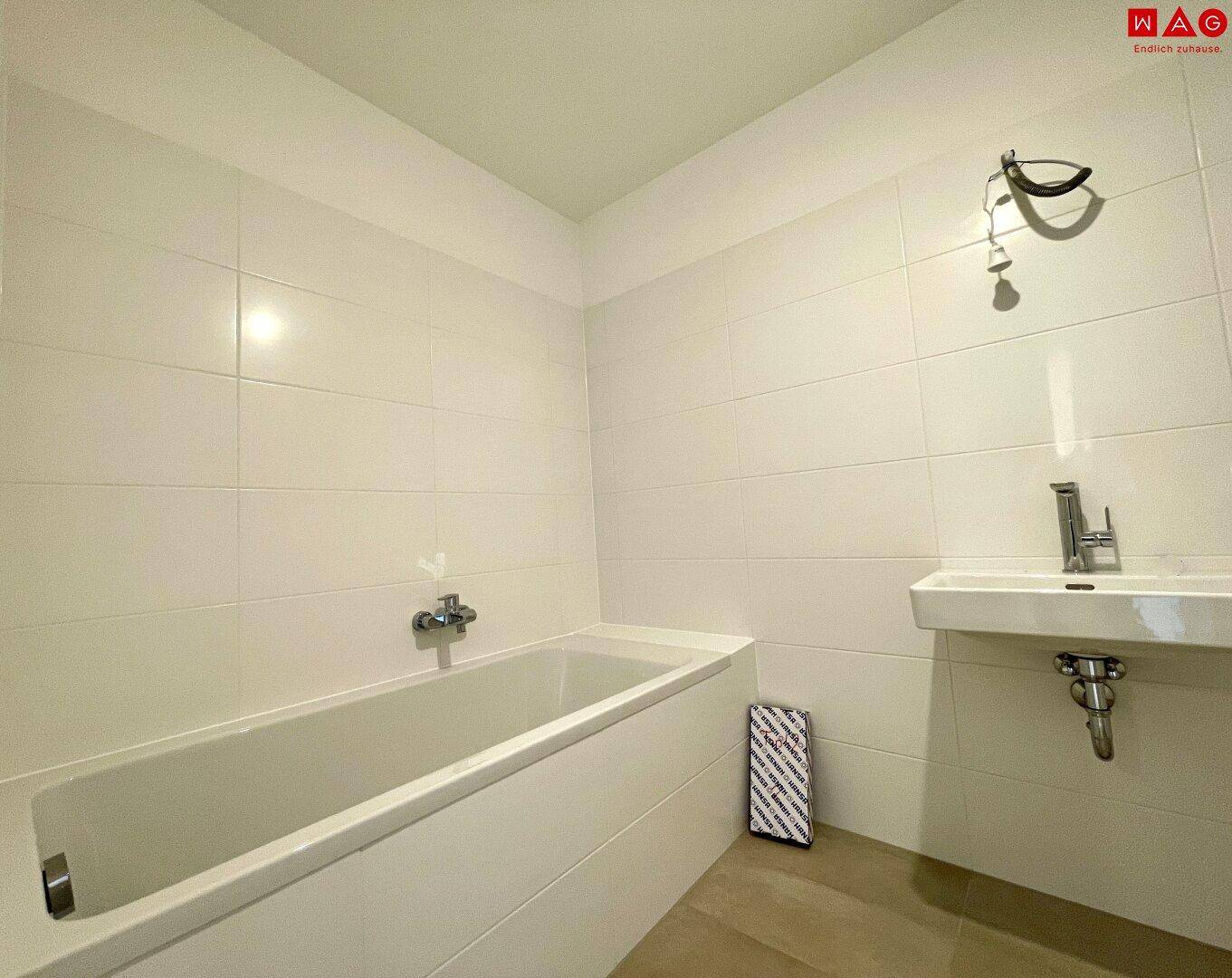 Helles Badezimmer mit Badewanne und kleinem Waschbecken, komplett gefliest.
