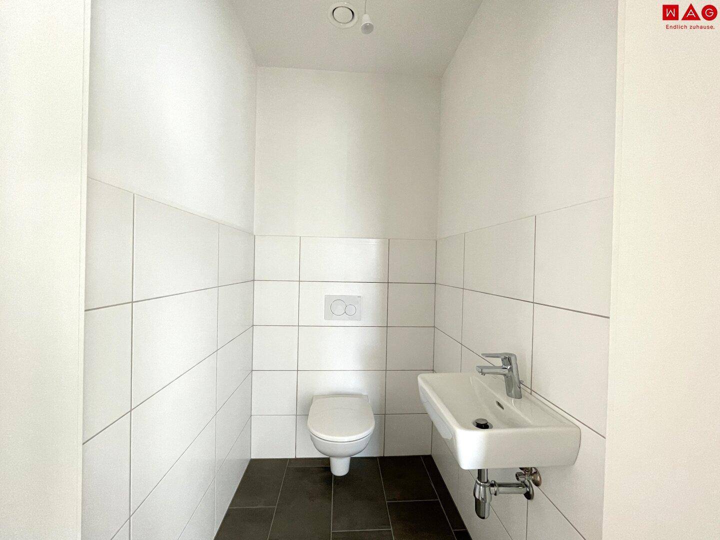 Helles Gäste-WC mit Toilette und Waschtisch, ausgestattet mit weißen Fliesen und dunklem Boden.