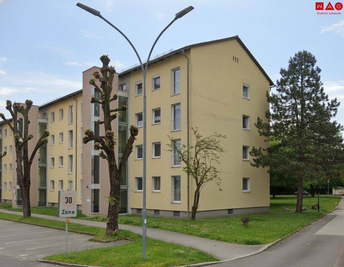 Modernes Mehrfamilienhaus mit beiger Fassade, vielen Fenstern und umgebender Grünfläche.