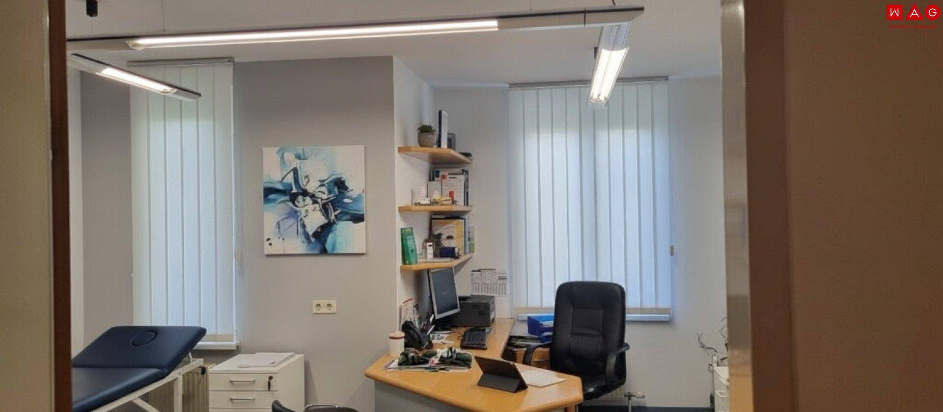 Helles Büro oder Sprechzimmer mit Schreibtisch, Bürostuhl und vertikalen Jalousien an den Fenstern.