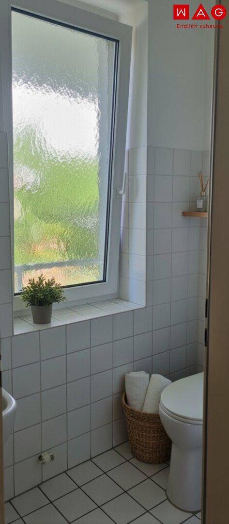 Kleines Badezimmer mit weißen Fliesen, Toilette und einem Fenster, dekoriert mit einer Topfpflanze.