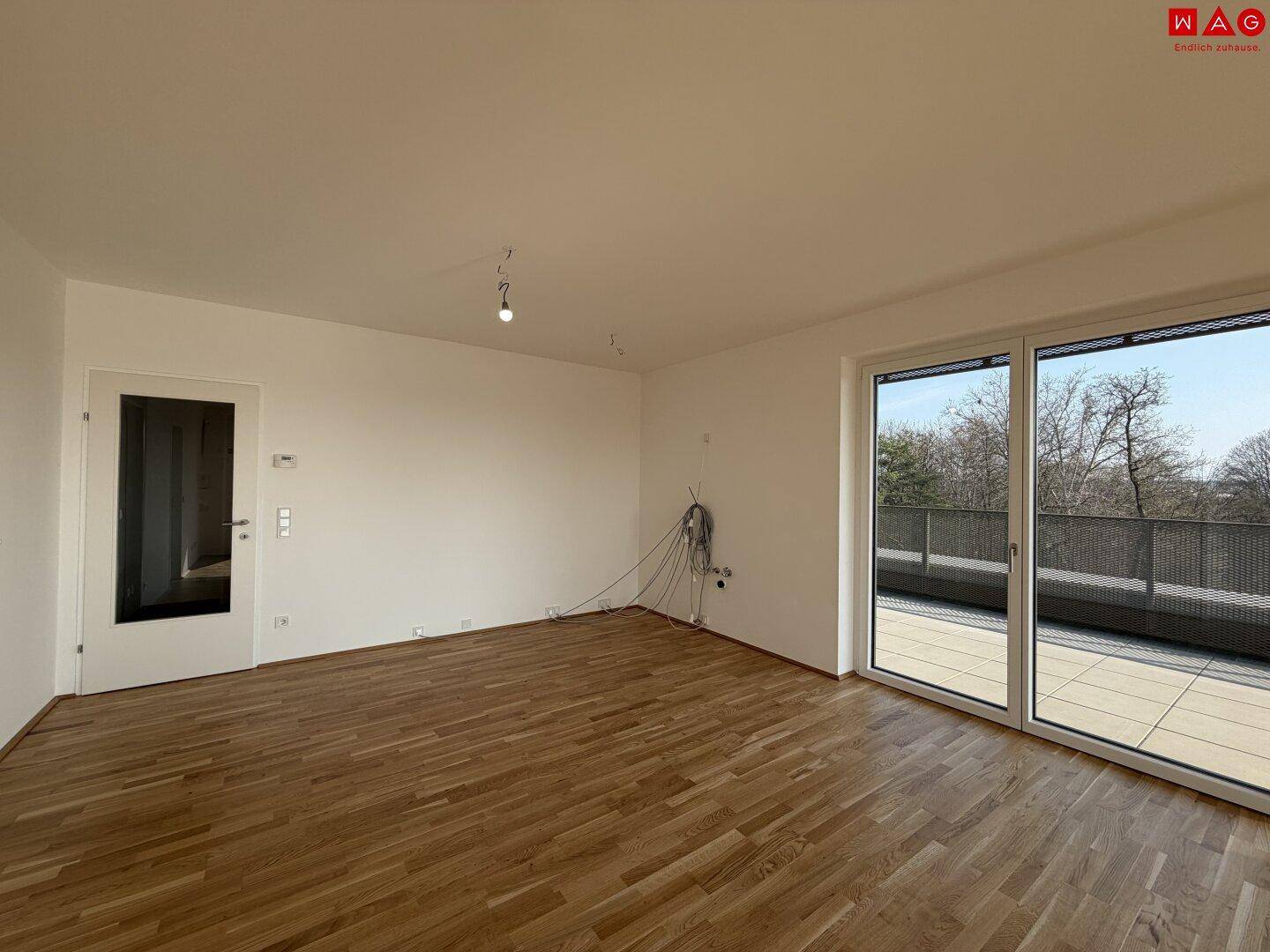 Geräumiger, unmöblierter Wohnbereich mit Holzboden und großen Fenstern, die auf den Balkon führen.