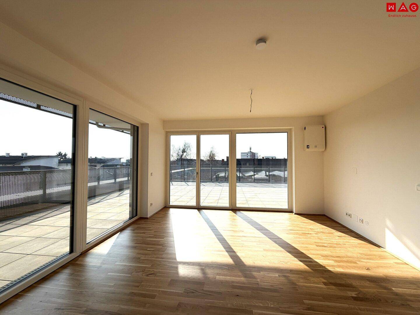 Heller, unmöblierter Wohnbereich mit Holzboden und großen Fenstern, die viel Tageslicht hereinlassen.
