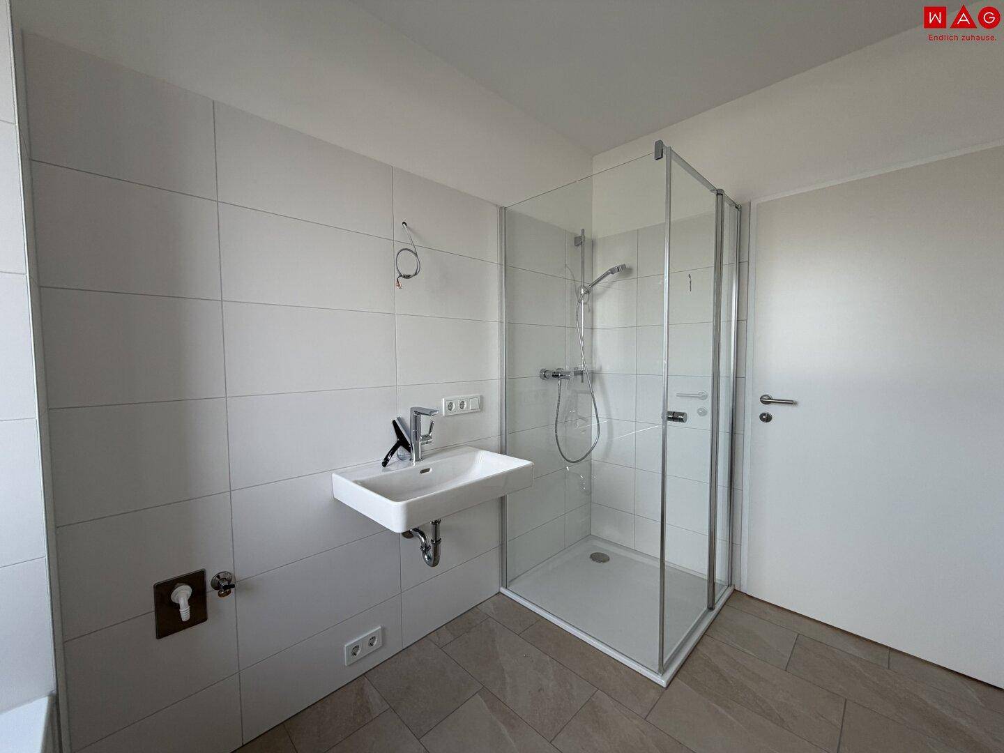 Modernes Badezimmer mit Duschkabine und Waschbecken, hell gefliest und mit viel Tageslicht.