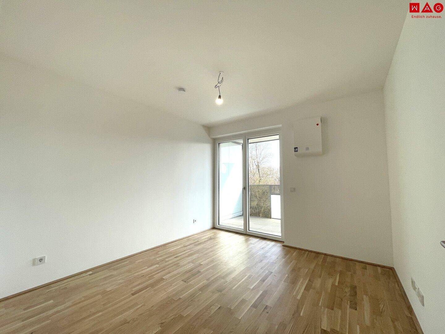 Leeres Schlafzimmer mit Parkettboden, Klimaanlage und Zugang zu einem sonnigen Außenbereich.