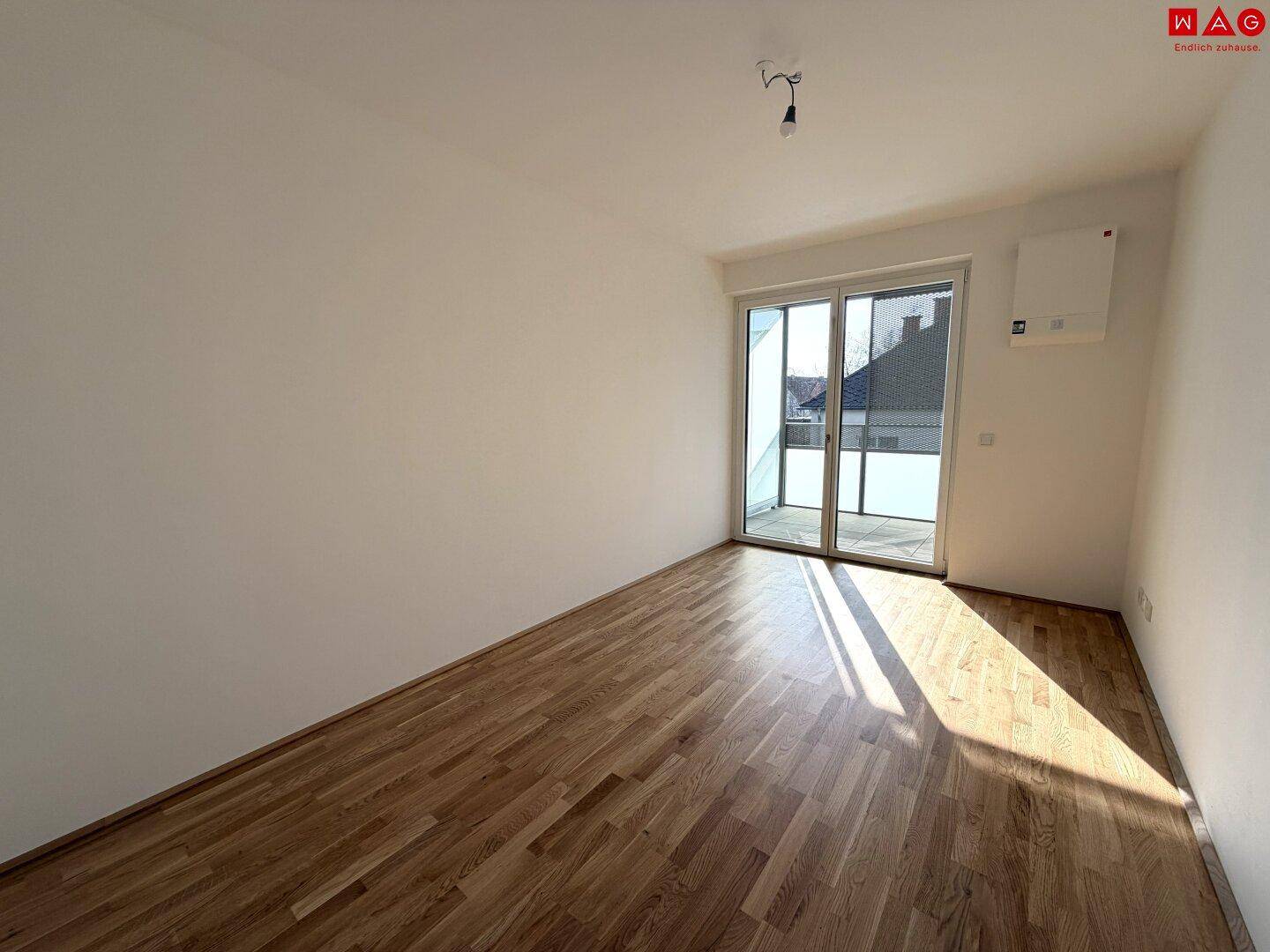 Leerer, sonniger Raum mit Parkettboden und Zugang zu einem Außenbereich.