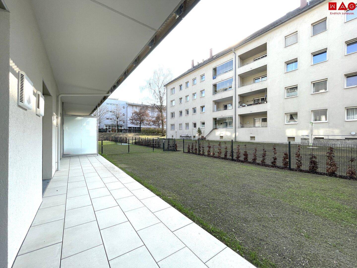 Lange, geflieste Terrasse mit Blick auf die grüne Rasenfläche und die umliegenden Wohngebäude.
