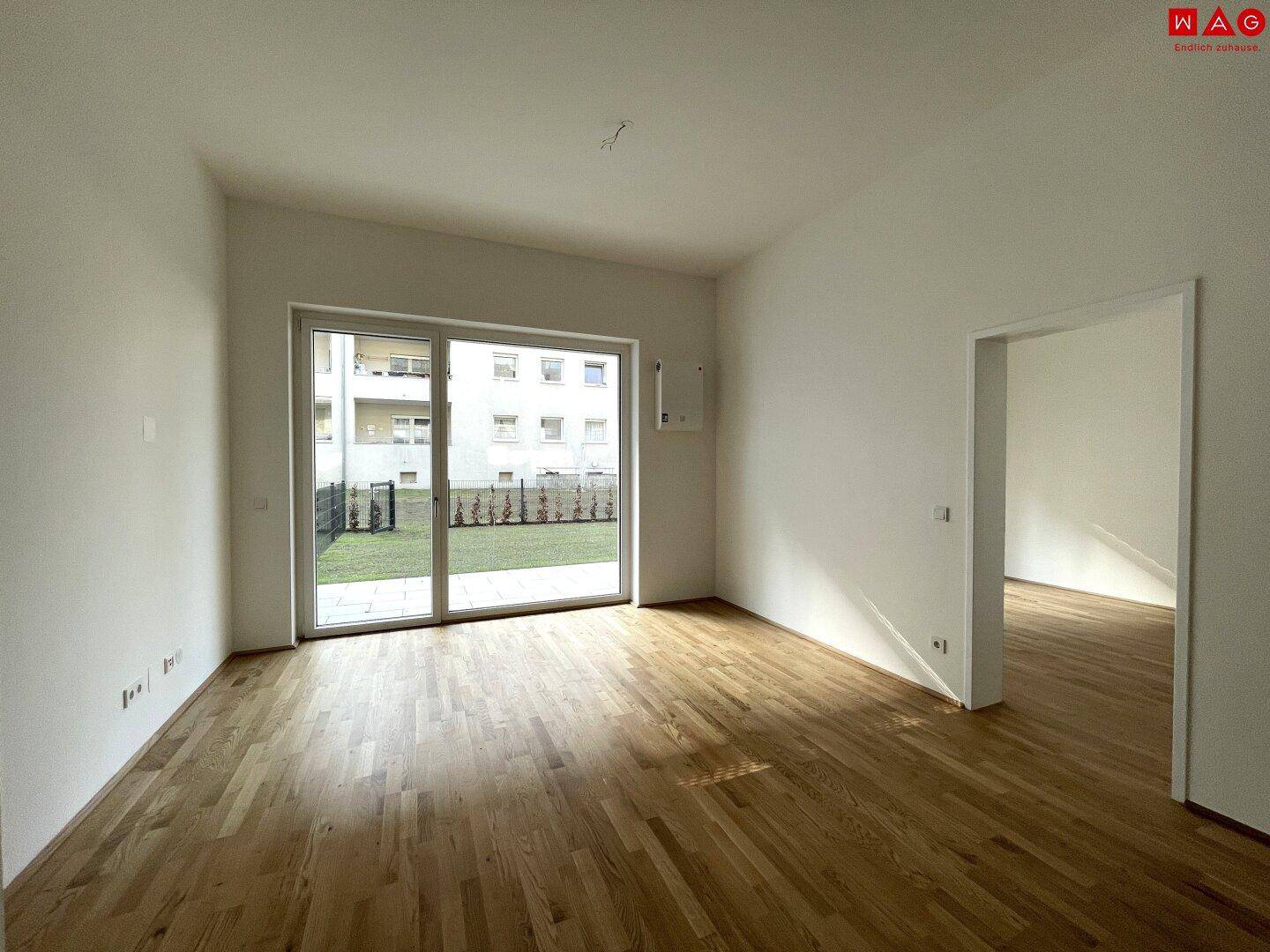 Leeres Wohnzimmer mit hellem Parkettboden und großem Fenster mit Blick auf die Grünfläche.