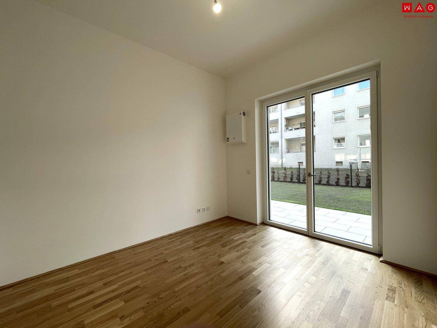 Leeres Zimmer mit Holzboden und großem Fenster, das viel Tageslicht hereinlässt.